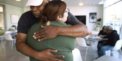 An older man in a white cap hugs a woman wearing glasses inside a bright room with tables and people in the background.