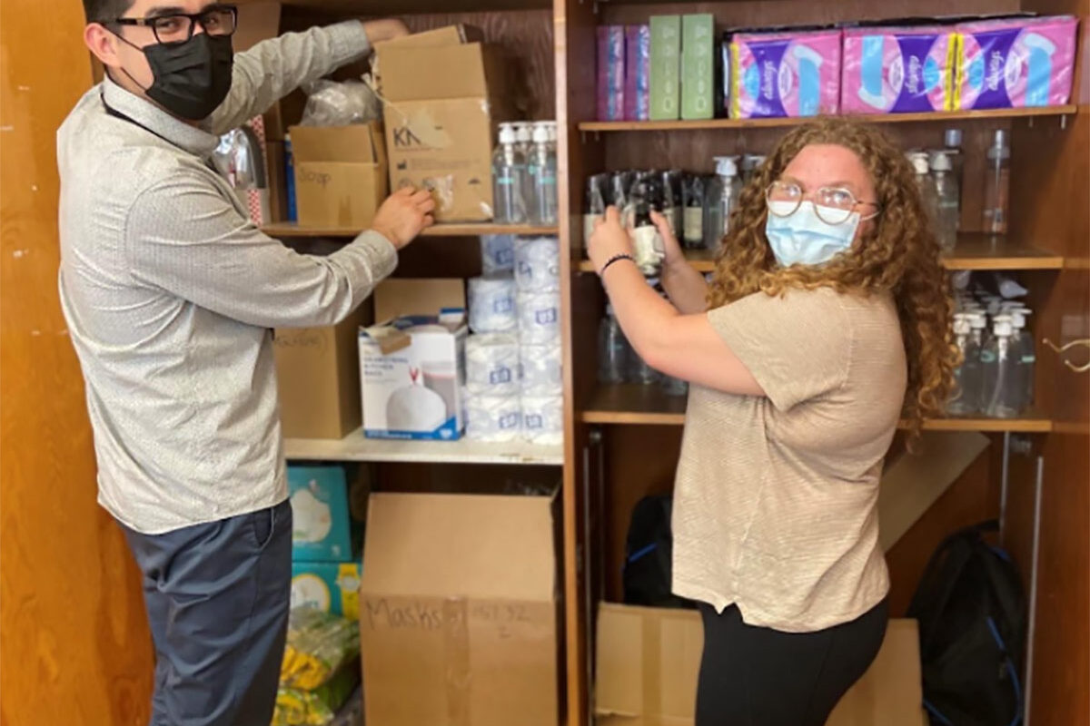 Two people wearing masks organize shelves stocked with sanitary products, bottled water, paper towels, and a box labeled “COVID-19 OUTBREAK: USE ONLY.”.