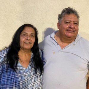 A woman and a man stand side by side, smiling, in front of a plain light-colored wall.