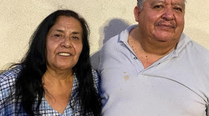 A woman and a man stand side by side, smiling, in front of a plain light-colored wall.