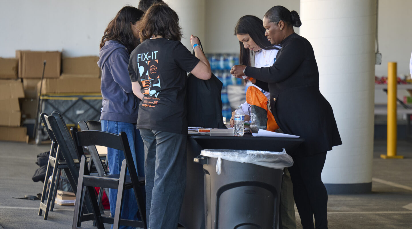 Five people stand around a table in a parking garage, distributing items with boxes in the background and a trash can in the foreground.