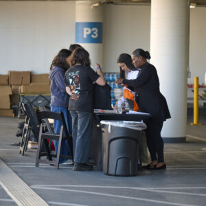 Five people stand around a table in a parking garage, distributing items with boxes in the background and a trash can in the foreground.