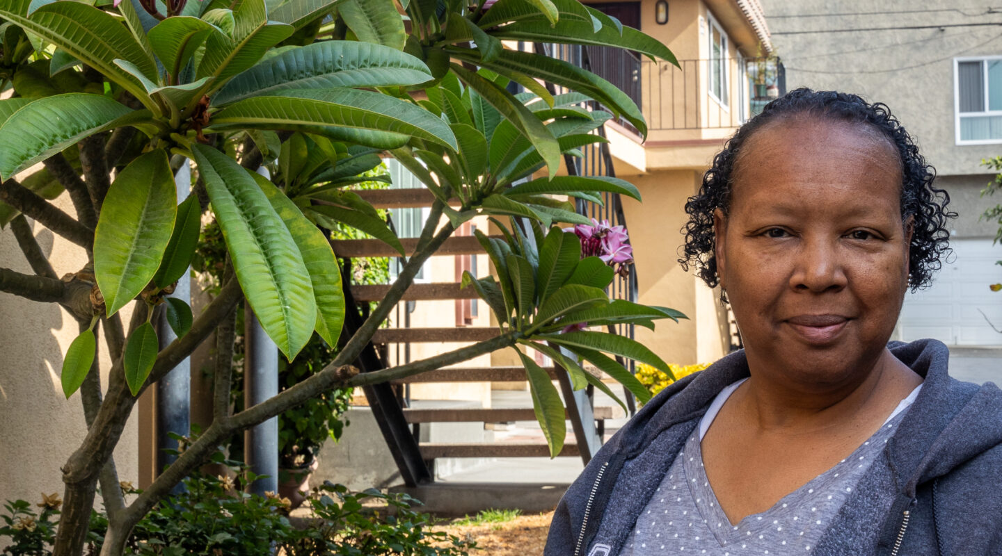 A woman stands beside a blooming plumeria tree in front of a residential building on a sunny day.