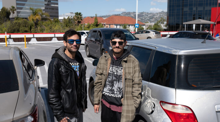 Two men wearing sunglasses stand next to a damaged car in a parking lot, with buildings and a hillside visible in the background.