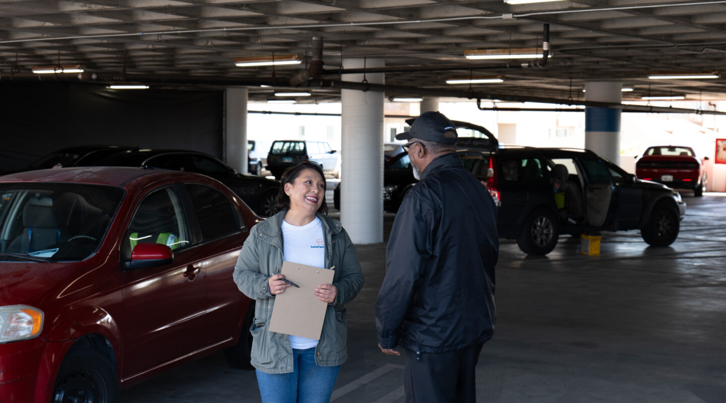 A woman holding a clipboard smiles while talking to a man in a hat inside a parking garage with several parked cars.