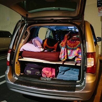 A minivan parked with its rear hatch open, showing a makeshift sleeping area with bedding, bags, and belongings organized on shelves inside.