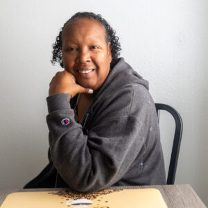 A person with short curly hair, wearing a gray zip-up hoodie, sits at a table, smiling and resting their chin on one hand.