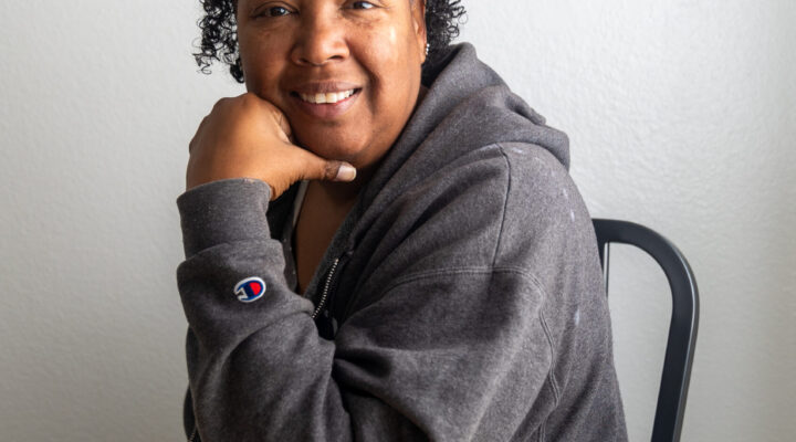 A person with short curly hair, wearing a gray zip-up hoodie, sits at a table, smiling and resting their chin on one hand.