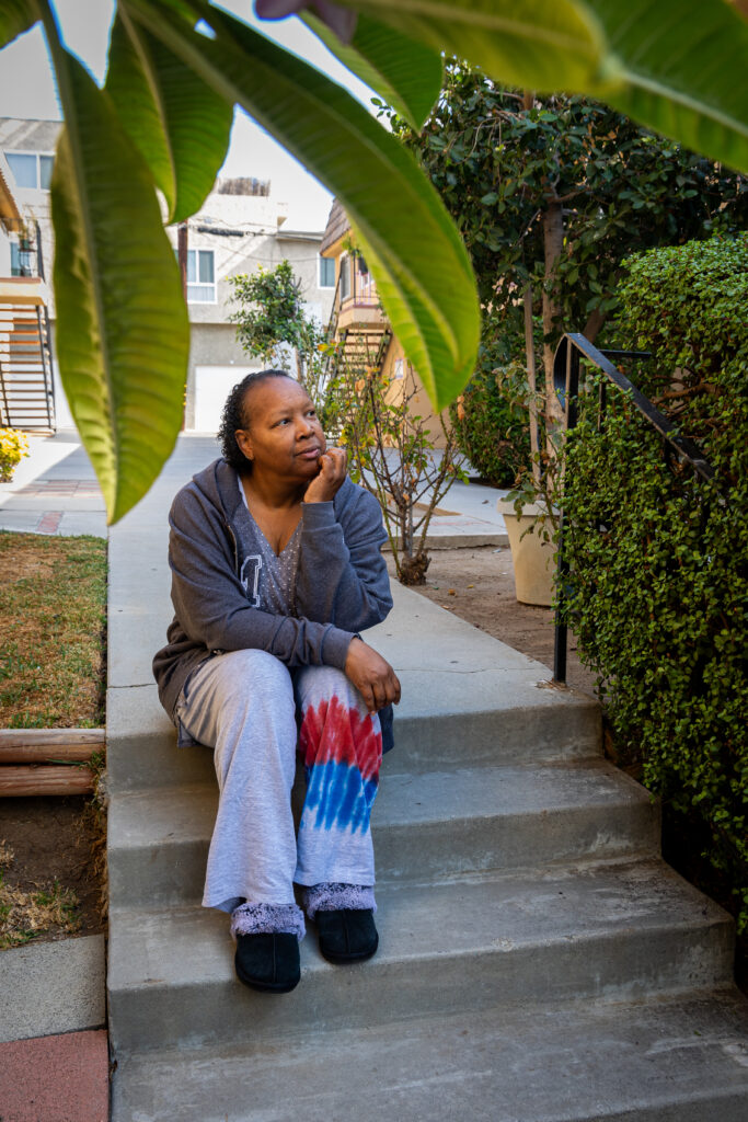A woman in casual clothes sits on outdoor steps, resting her chin on her hand, with greenery and apartment buildings in the background.