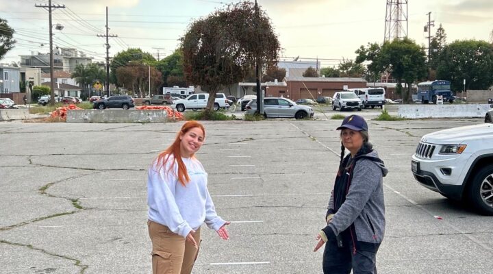 Two people stand in an empty parking lot, each pointing down at lines marked on the ground. Cars and utility poles are visible in the background under a cloudy sky.