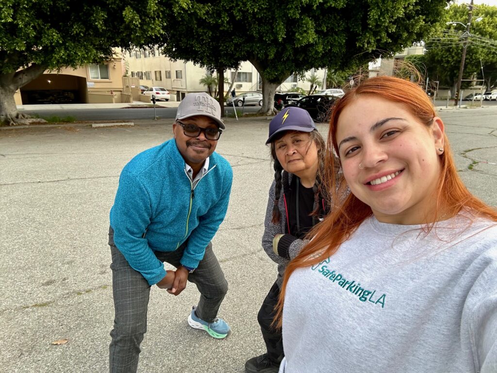 Three people pose together outdoors in a parking lot, with trees and buildings in the background.