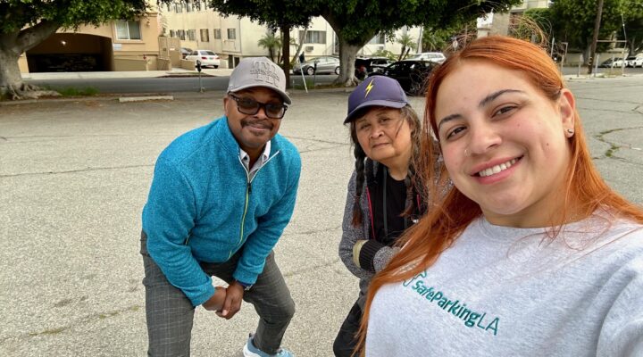 Three people pose together outdoors in a parking lot, with trees and buildings in the background.