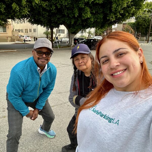 Three people pose for a selfie in an outdoor parking lot; trees and parked cars are visible in the background.