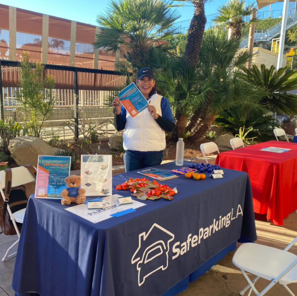 A woman stands behind a Safe Parking LA table with brochures, snacks, and informational materials displayed at an outdoor event.