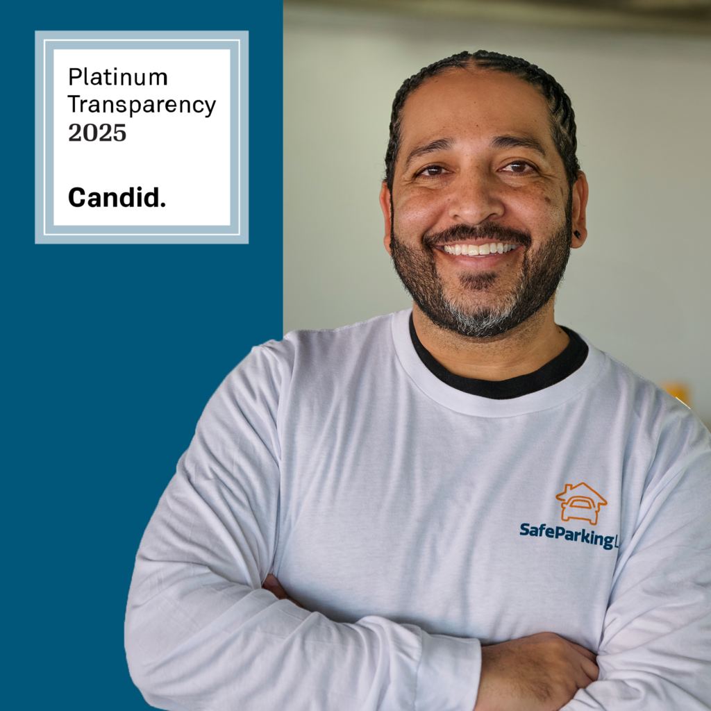 Smiling man with folded arms wearing a SafeParkingLA shirt stands next to a sign reading “Platinum Transparency 2025 Candid.”.