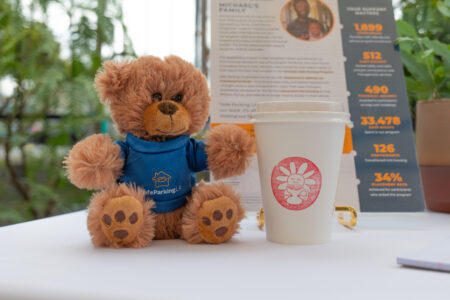 A brown teddy bear in a blue shirt sits next to a white coffee cup with a pink sun logo on a table, with informational pamphlets in the background.
