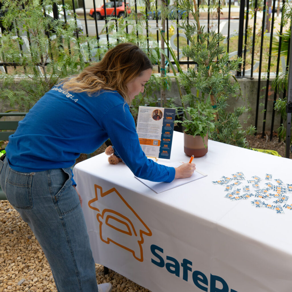 A woman in a blue shirt leans over a table, writing on a sheet of paper at an outdoor SafePlace event booth. The table holds flyers and a potted plant.