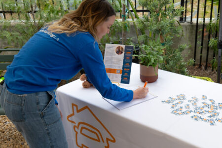 A woman in a blue shirt leans over a table, writing on a sheet of paper at an outdoor SafePlace event booth. The table holds flyers and a potted plant.