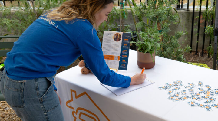 A woman in a blue shirt leans over a table, writing on a sheet of paper at an outdoor SafePlace event booth. The table holds flyers and a potted plant.
