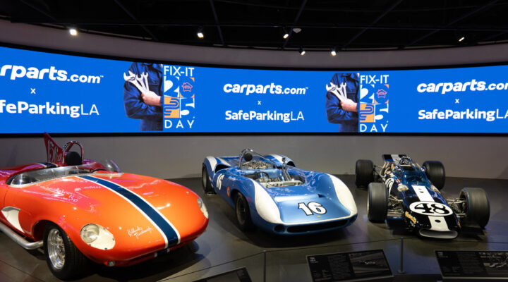Three vintage race cars are displayed in a museum with informational plaques in front and a large digital screen in the background featuring carparts.com and SafeParkingLA logos.