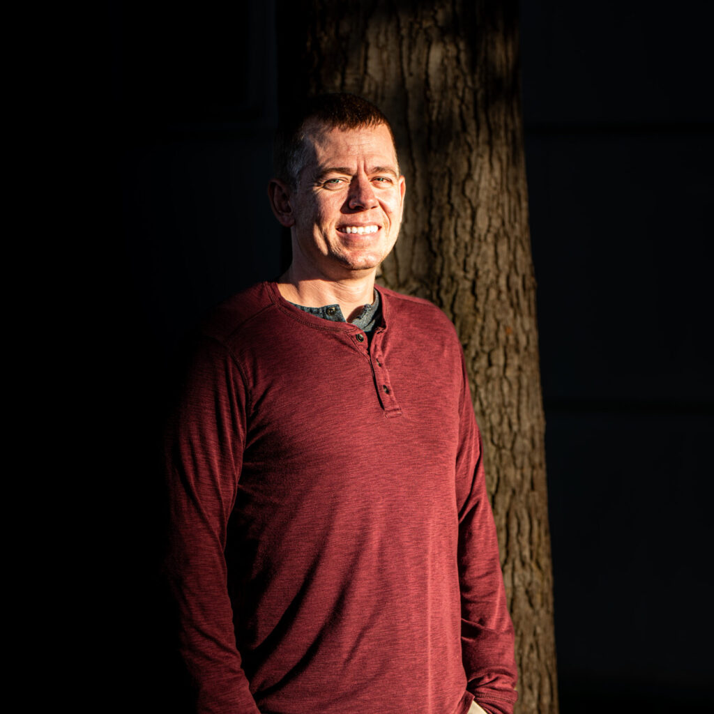 A man in a maroon long-sleeve shirt stands in sunlight next to a tree, smiling, with a dark background.