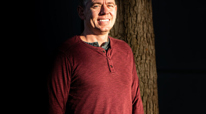 A man in a maroon long-sleeve shirt stands in sunlight next to a tree, smiling, with a dark background.