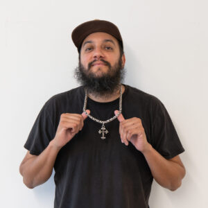 A man with a beard and cap stands against a white background, holding the chain of his necklace with both hands while wearing a black t-shirt.