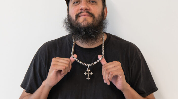 A man with a beard and cap stands against a white background, holding the chain of his necklace with both hands while wearing a black t-shirt.