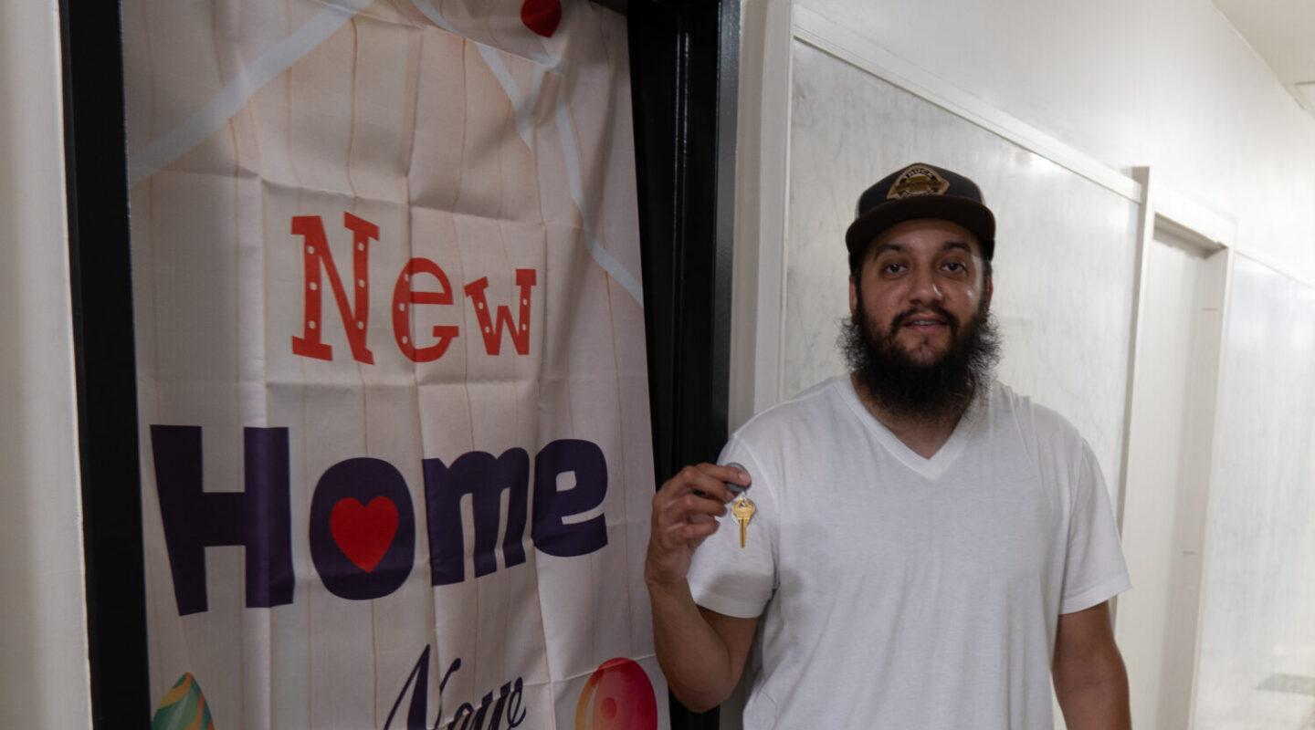 A man stands in a hallway holding a key and smiling in front of a door with a