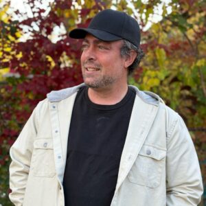 A man wearing a black cap, black shirt, and light-colored jacket stands outdoors in front of autumn foliage, looking to the side and smiling.