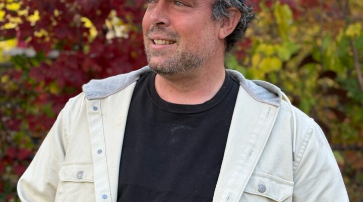 A man wearing a black cap, black shirt, and light-colored jacket stands outdoors in front of autumn foliage, looking to the side and smiling.