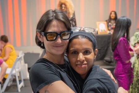 Two people wearing black shirts and glasses hug and smile at the camera in a brightly lit indoor setting. Both are proud to volunteer with Safe Parking LA, with others and a table visible in the background.