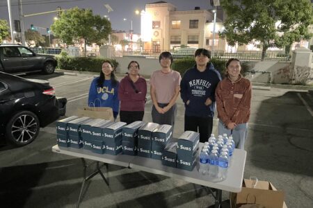 Five people volunteer with Safe Parking LA, standing behind a table with sandwich boxes and water bottles in a parking lot at night, with buildings and trees in the background.
