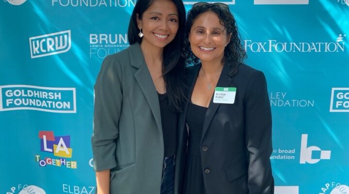 Two women stand side by side, smiling at the camera, in front of a blue step-and-repeat banner displaying multiple sponsorship logos.