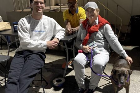 Three people sit at outdoor tables at night; one woman, a volunteer with Safe Parking LA, holds a dog on a leash. The setting appears casual and relaxed.