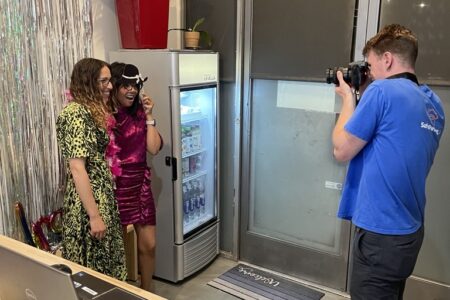 A photographer captures two people posing and smiling in front of a decorated backdrop next to a refrigerator, highlighting their experience as they volunteer with Safe Parking LA in an indoor setting.
