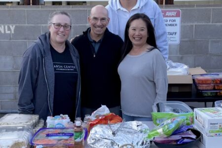 Four people, volunteer with Safe Parking LA, stand behind a table with various food items and drinks, smiling at the camera outdoors near a brick wall and a reserved parking sign.