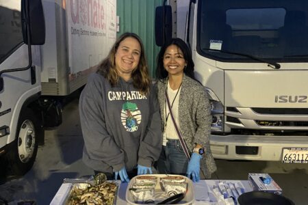Two women volunteer with Safe Parking LA, standing behind a table with snacks and supplies, smiling at the camera, as trucks and a 