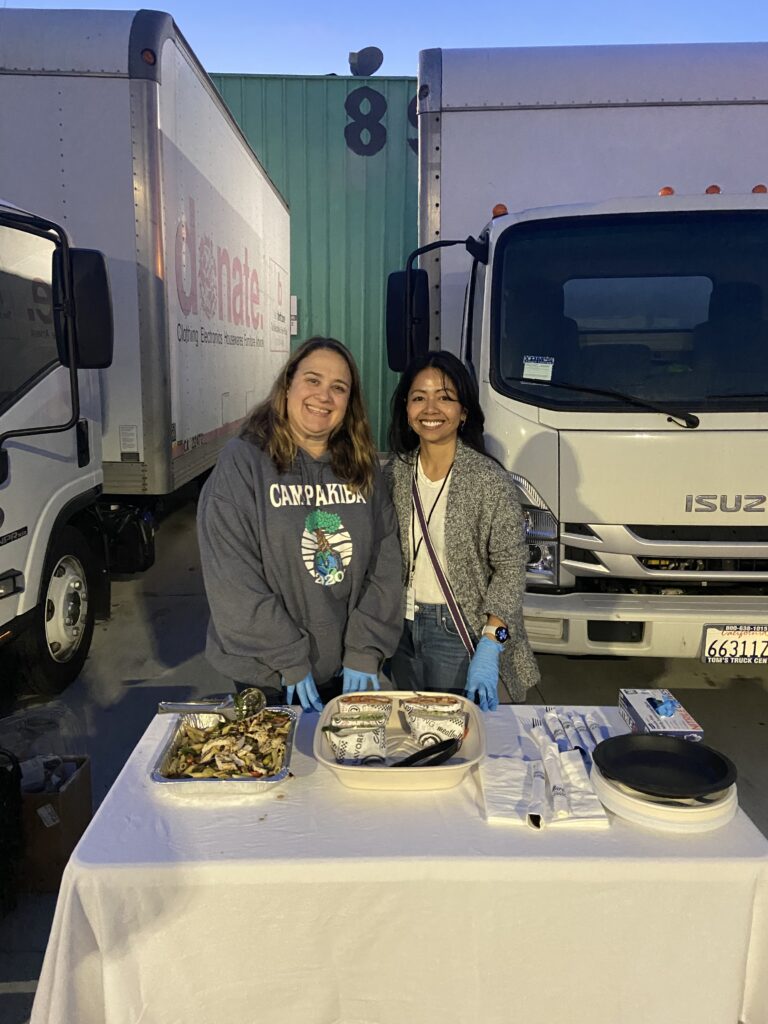 Two women volunteer with Safe Parking LA, standing behind a table with snacks and supplies, smiling at the camera, as trucks and a "donate" sign appear in the background.