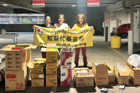 Three people, who volunteer with Safe Parking LA, hold a SimplyHelp Foundation banner in a parking garage, standing behind boxes of donated food and supplies.