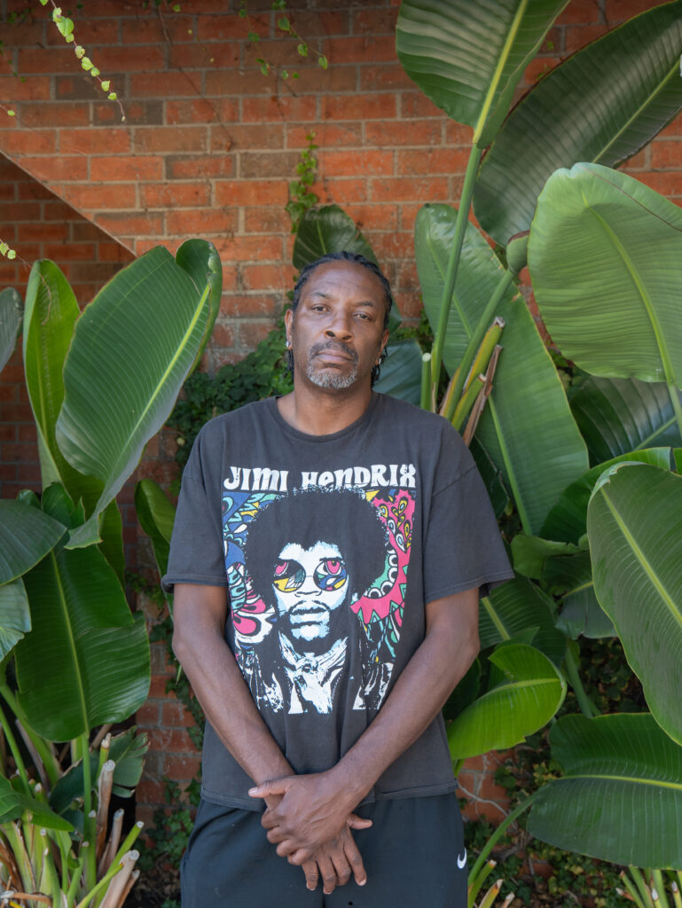 A man stands in front of large green plants and a brick wall, wearing a Jimi Hendrix t-shirt and dark pants, with his hands clasped in front of him.