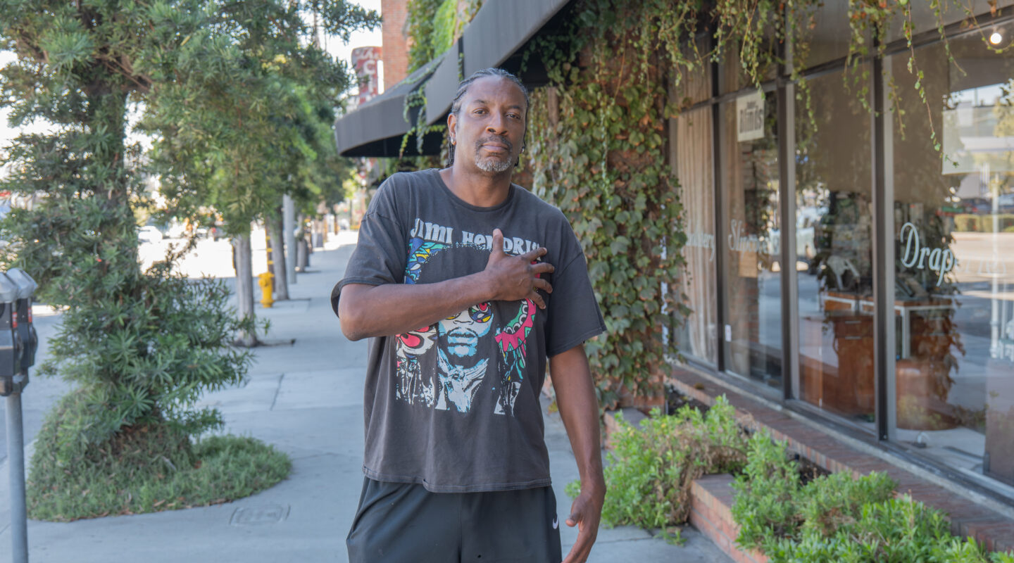 A man stands on a sidewalk near a building covered with ivy, wearing a graphic T-shirt and shorts, with one hand touching his chest.