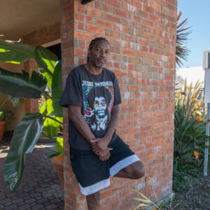 A man in a Jimi Hendrix t-shirt and shorts leans against a brick wall outside, with plants and a parking lot in the background.