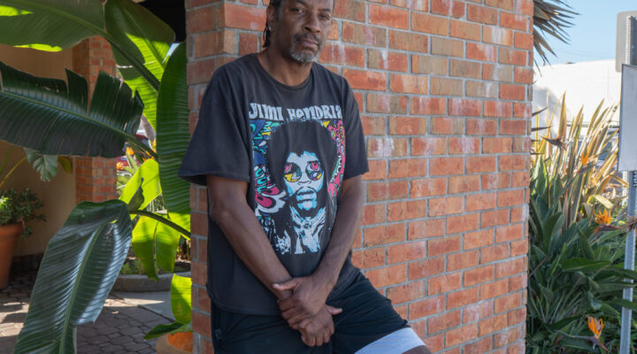 A man in a Jimi Hendrix t-shirt and shorts leans against a brick wall outside, with plants and a parking lot in the background.
