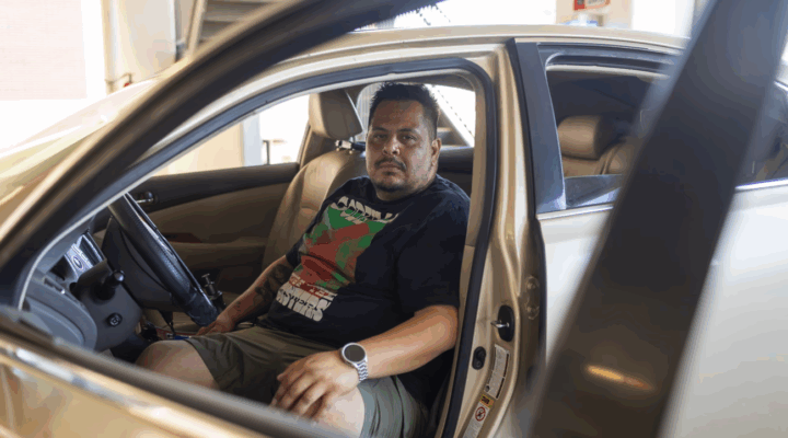 A man sits in the driver's seat of a beige car with the door open, looking ahead.