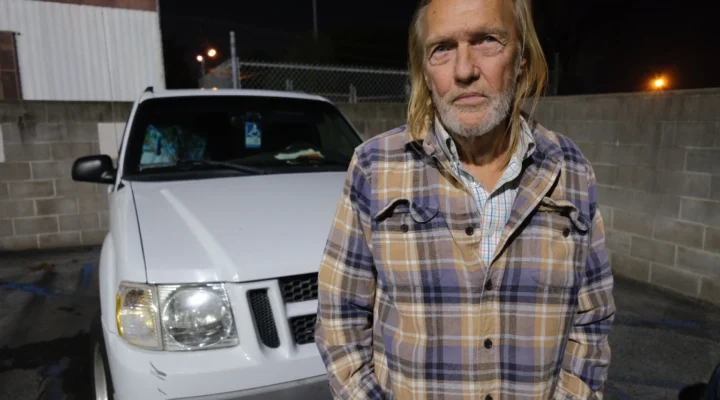 An older man in a plaid shirt stands in front of a white pickup truck in a parking lot at night.