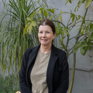 A woman stands outside in front of green plants, wearing a black jacket, beige shirt, and blue jeans, with a black bag over her shoulder.