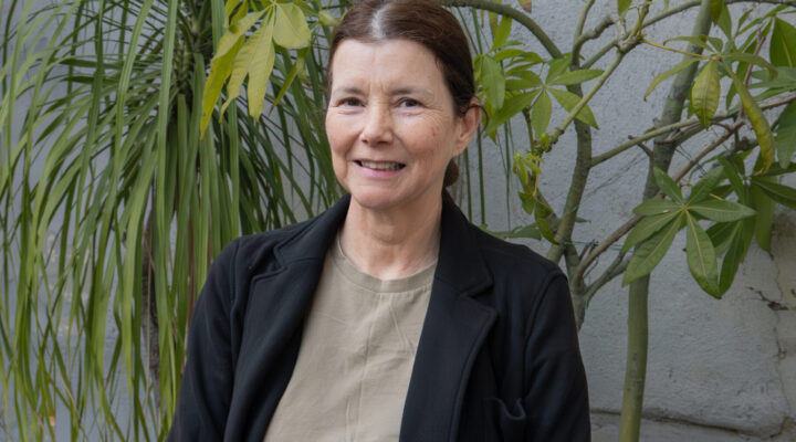 A woman stands outside in front of green plants, wearing a black jacket, beige shirt, and blue jeans, with a black bag over her shoulder.