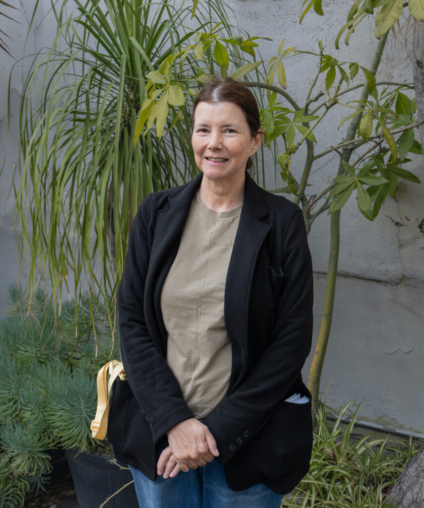 A woman stands outdoors in front of green plants, wearing a black jacket, beige shirt, and blue jeans, with a black bag over her shoulder.