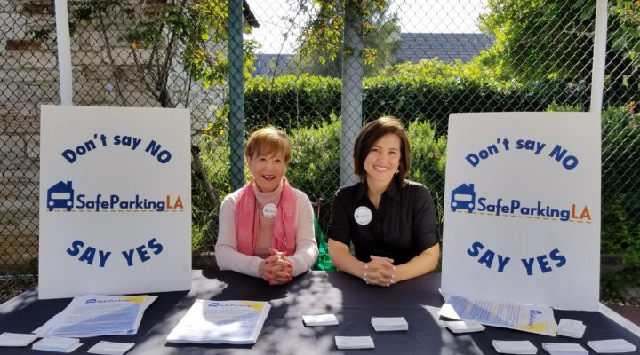 Two women sit at a table under a canopy with “Safe Parking LA” signs, pamphlets, and business cards at a community outreach event.